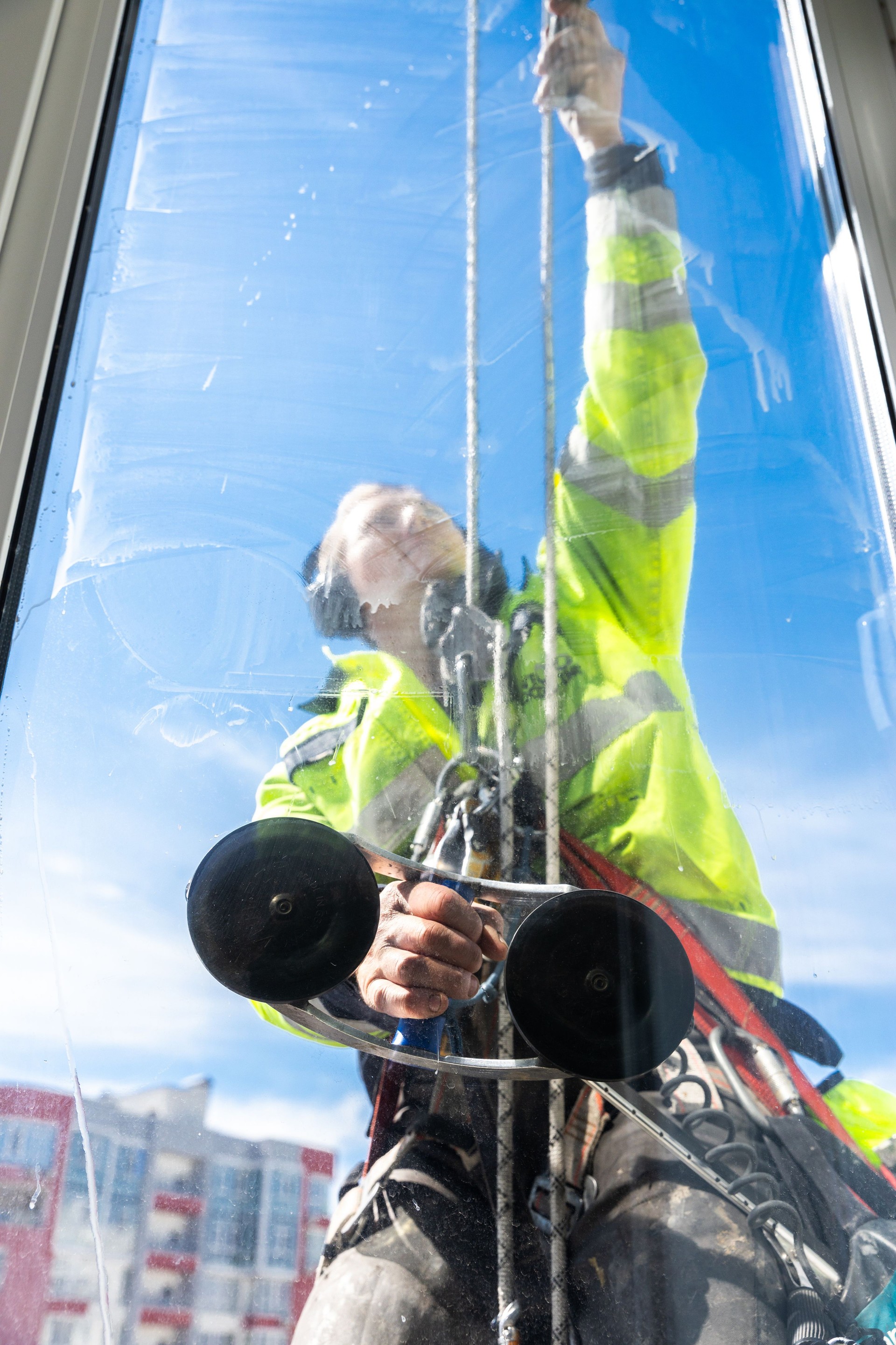Professional Window Cleaner Man Cleans High-Rise Building Windows with Safety Harness and Equipment in Bright Blue Sky Environment